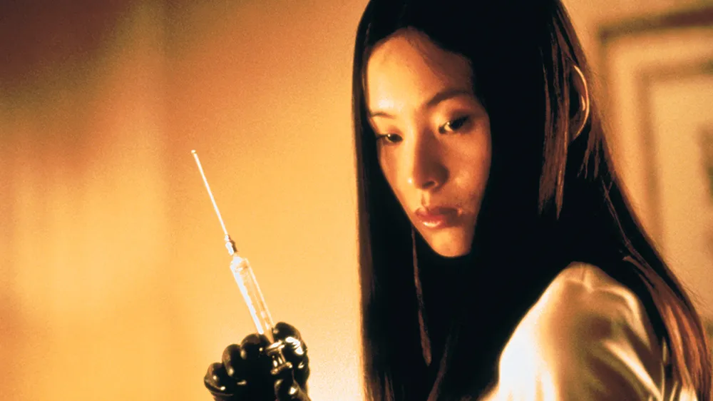 A chilling scene from the 1999 horror film Audition, featuring Asami Yamazaki, played by Eihi Shiina, holding a syringe with a calm yet unsettling expression. Wearing black gloves and a white dress, she exudes quiet menace against a warm, dimly lit background, capturing the film’s eerie and suspenseful atmosphere.