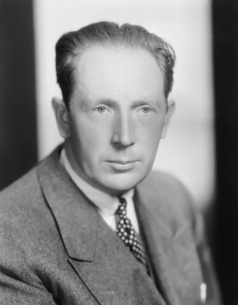 Black and white portrait of F.W. Murnau, the German filmmaker behind Nosferatu (1922). He is dressed in a suit with a patterned tie, his neatly combed hair and serious expression reflecting his visionary presence in early cinema. The soft lighting and blurred background emphasize his intense gaze and artistic demeanor.