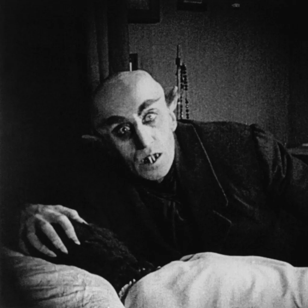 Max Schreck as Count Orlok in Nosferatu (1922), looming over a sleeping victim in a chilling black and white scene. His gaunt, bald head, pointed ears, and wide, menacing eyes add to his eerie, undead presence. His clawed hand rests on the victim, while his fanged mouth is slightly open, frozen in a moment of silent horror.