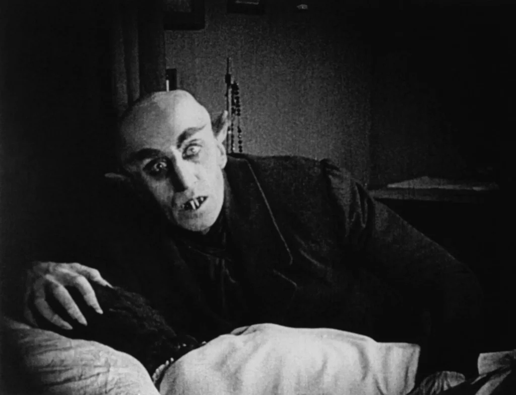Black and white still from Nosferatu showing the vampire with a bald head, pointed ears, sharp teeth, and claw-like fingers leaning over a sleeping woman.
